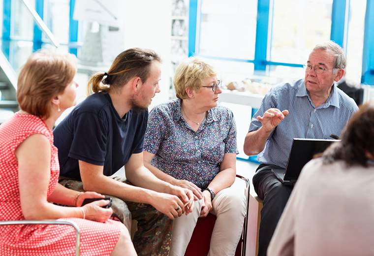 A group of four people sit in a well-lit room engaged in conversation. An older man gestures while speaking, evoking a collaborative and attentive mood.