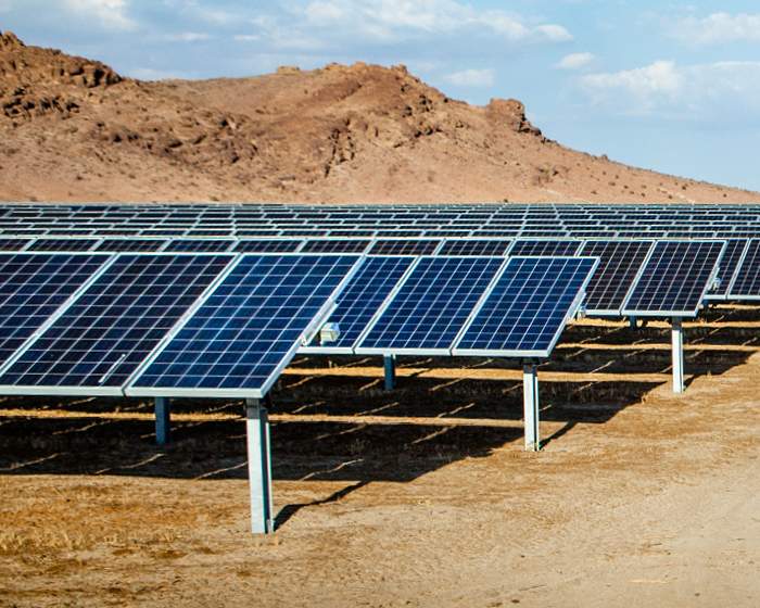 Rows of solar panels stretch across a desert landscape under a blue sky with scattered clouds, conveying a sense of clean energy and sustainability.