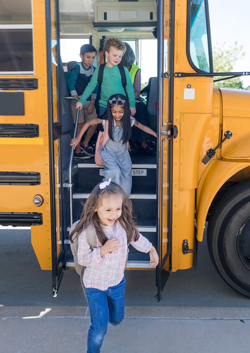 Children excitedly exit a yellow school bus. A girl in the foreground runs with a smile, while others behind her step down, conveying a joyful school day.
