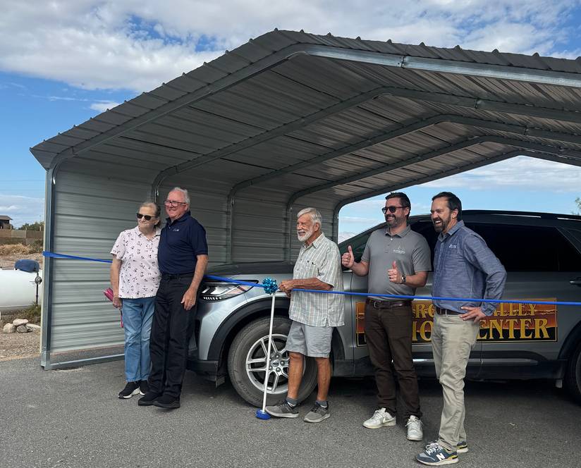 Nye county residents stand smiling under a metal carport with a vehicle in the background. A blue ribbon is stretched in front, conveying a celebratory event.
